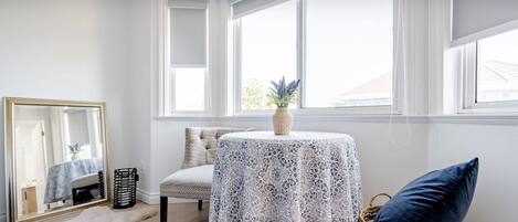 Ein Zimmer mit einem großen Fenster, einem Tisch mit weißer Spitze, einem Stuhl, einem Spiegel und einem blauen Kissen auf einem Sockel.