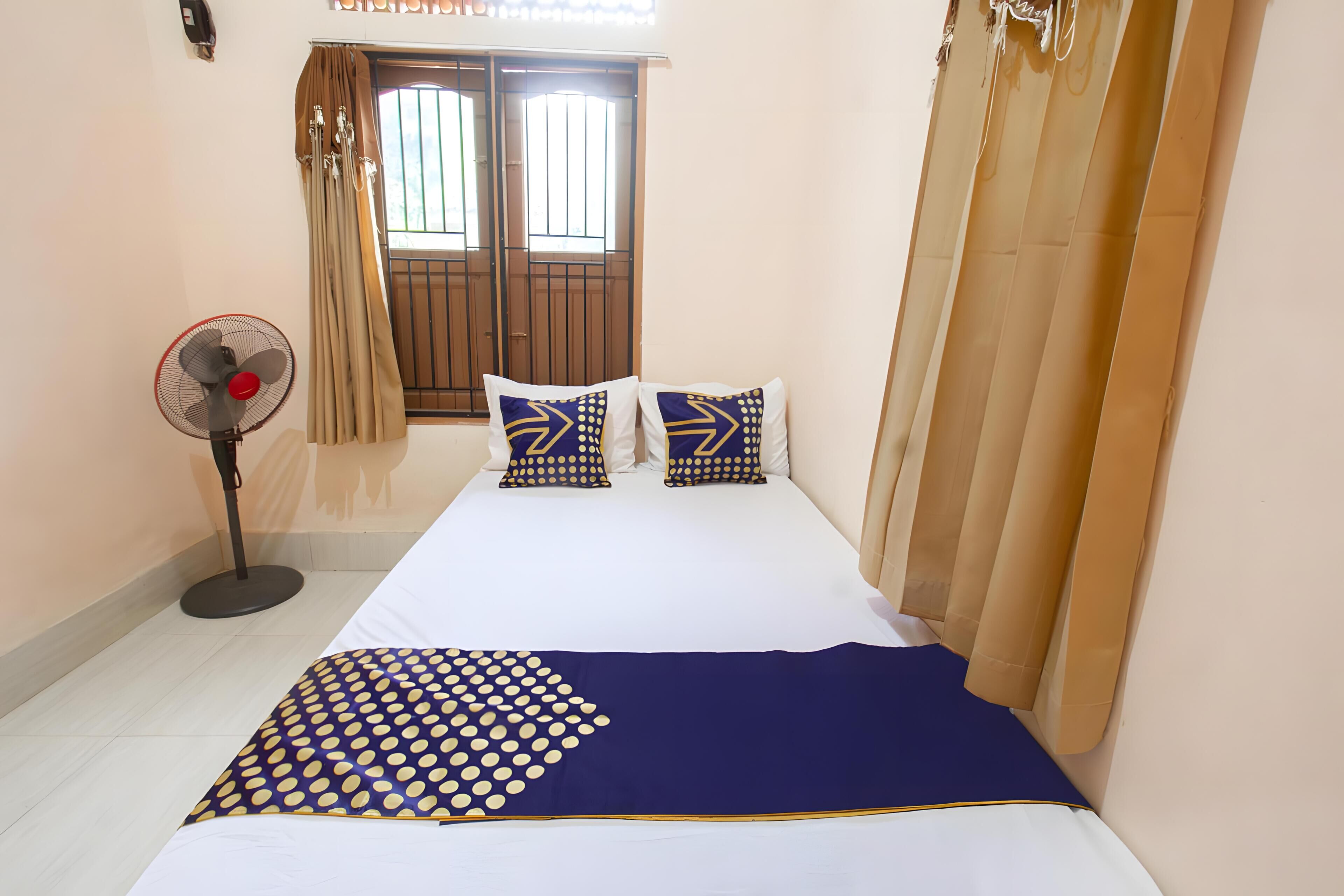 A bedroom with a bed, two pillows, a standing fan, and a window with curtains.