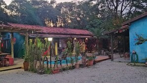 A rustic outdoor area with a pool, wooden tables, and chairs, surrounded by potted plants and a mural on the wall. - Recanto Zen (Imbituba)