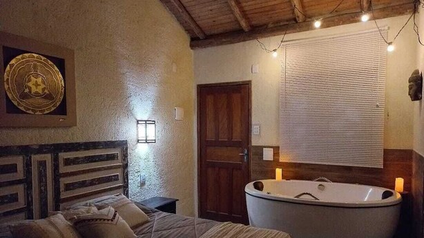 A bedroom with a large bed, a wooden headboard, a wall-mounted light, a wooden door, and a freestanding bathtub. - Recanto Zen (Imbituba)