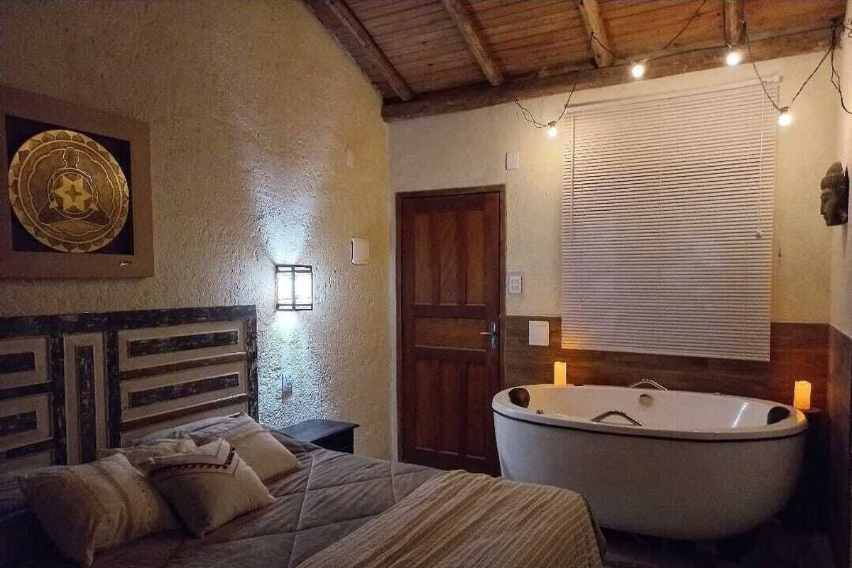 A bedroom with a large bed, a wooden headboard, a wall-mounted light, a wooden door, and a freestanding bathtub.