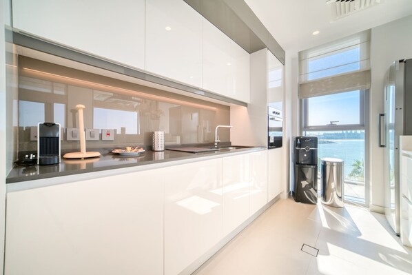 A modern kitchen with sleek white cabinetry, a stainless steel range hood, and a view of the sea through large windows.