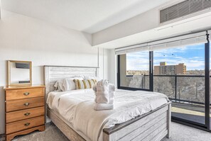A bedroom with a large window, a wooden dresser, a bed with a white comforter, and a view of buildings outside. - Mid-Nest Unit 708 (Colorado Springs)