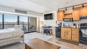 A modern hotel room with a bed, a dining table, a kitchen area with wooden cabinets, a flat-screen TV, and a city view through large windows. - Mid-Nest Unit 708 (Colorado Springs)