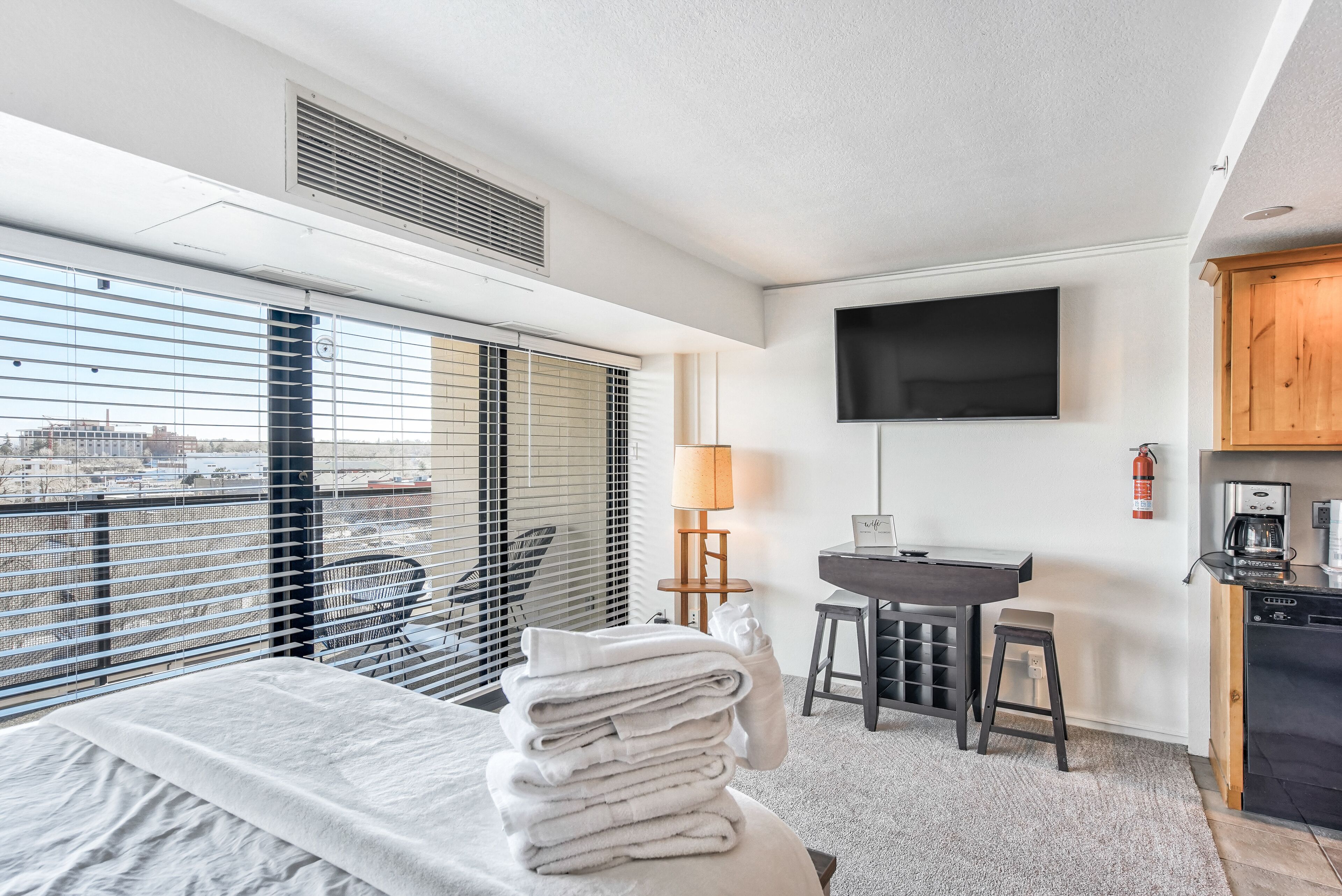 A hotel room with a bed, a desk, a chair, a TV, and a kitchen area.