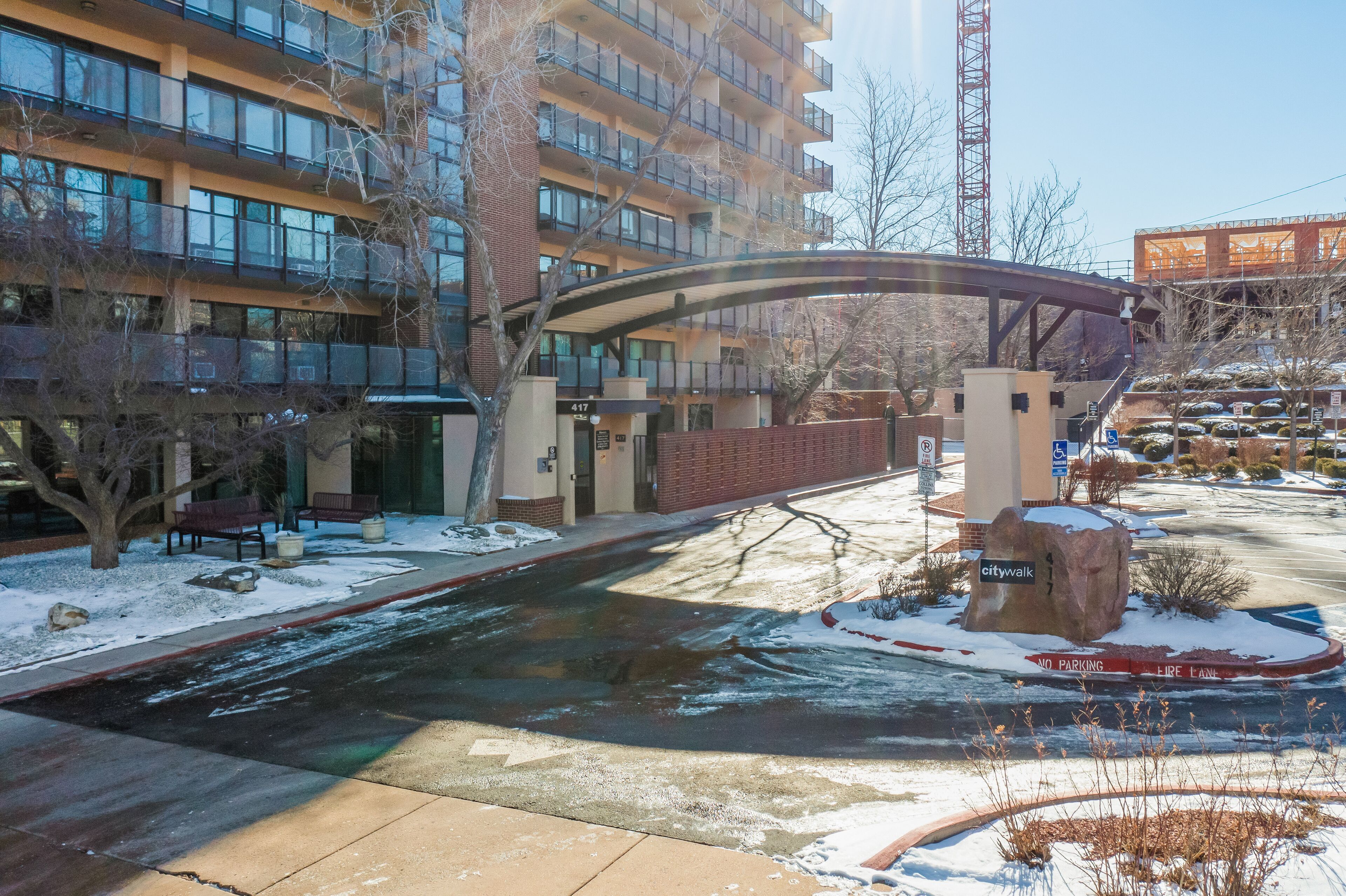 A multi-story building with a covered pedestrian bridge, snow on the ground, and a parking area with a 'No Parking' sign.