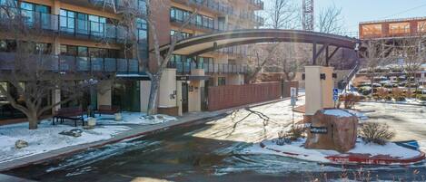A multi-story building with a covered pedestrian bridge, snow on the ground, and a parking area with a 'No Parking' sign.