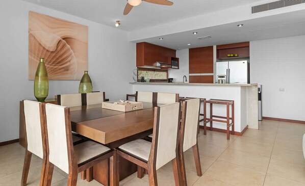 A dining area with a wooden table and chairs, a kitchen in the background, and a ceiling fan.