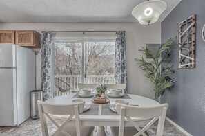 A kitchen with a dining table set for two, a refrigerator, and a window with curtains.