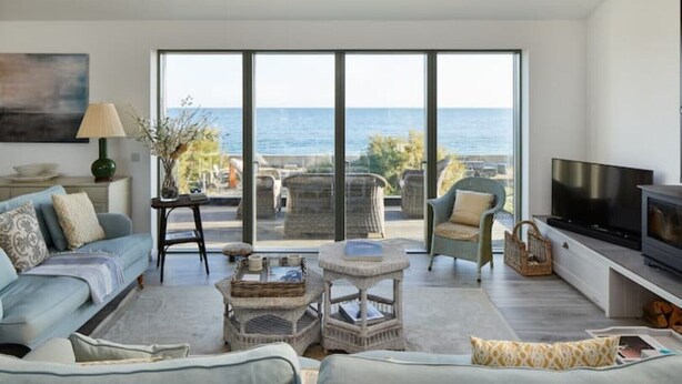 A modern living room with a large sliding glass door leading to a balcony with a view of the sea. - Stunning architect designed eco beachfront property (Selsey)