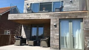 A modern two-story building with a wooden facade, large windows, and a covered patio area with outdoor furniture. - Stunning architect designed eco beachfront property (Selsey)