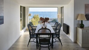 A dining area with a table and chairs, a vase with flowers, and a view of the sea through large windows. - Stunning architect designed eco beachfront property (Selsey)