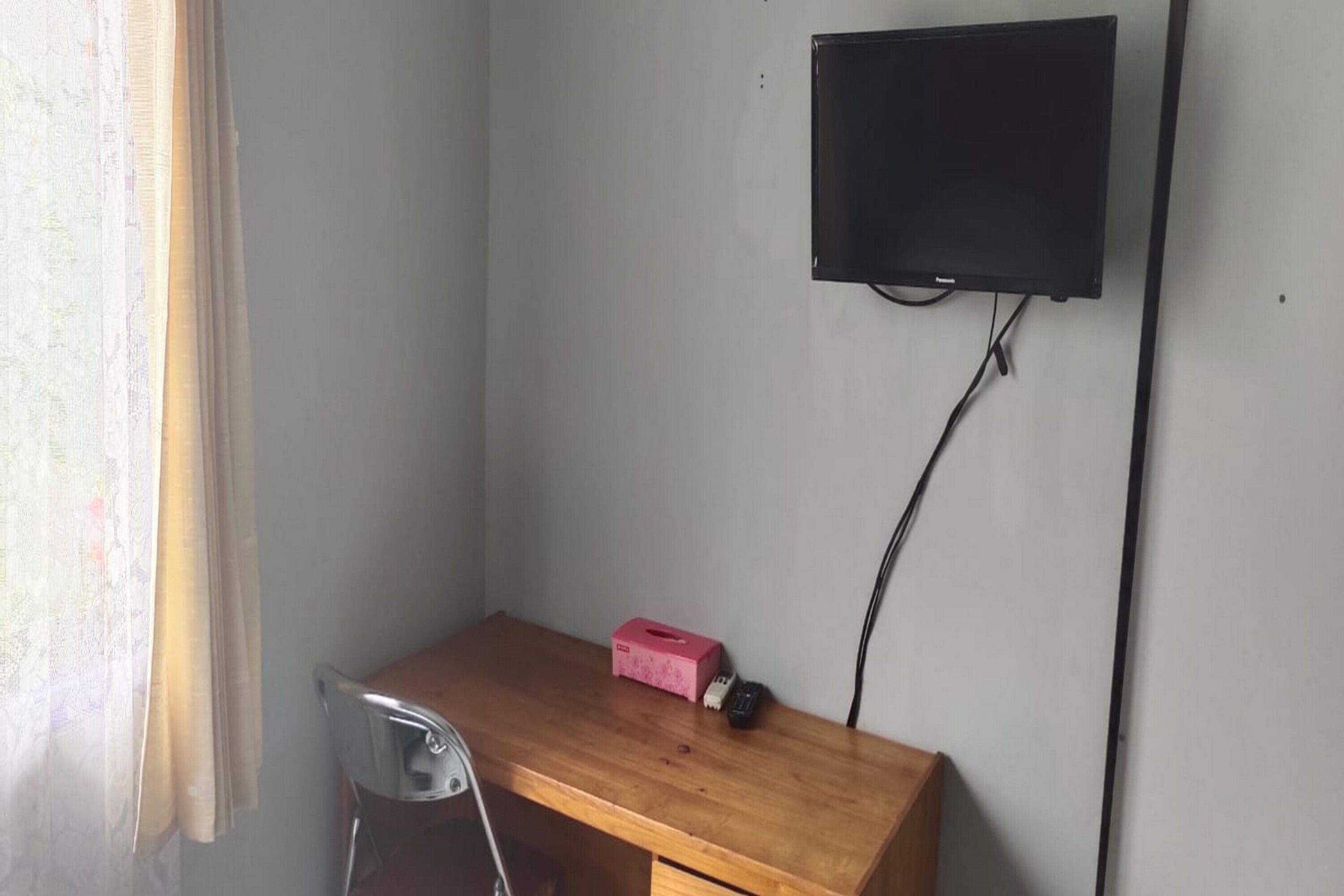 A small room with a wooden desk, a chair, a television mounted on the wall, and a window with curtains.