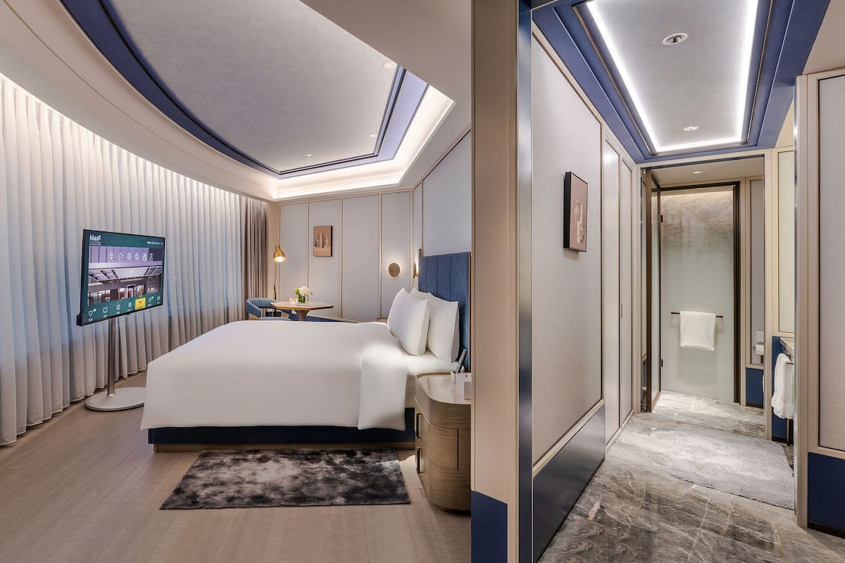 Una habitación de hotel moderna con una cama grande, un televisor de pantalla plana, una mesita con un florero y un baño con puerta de vidrio.