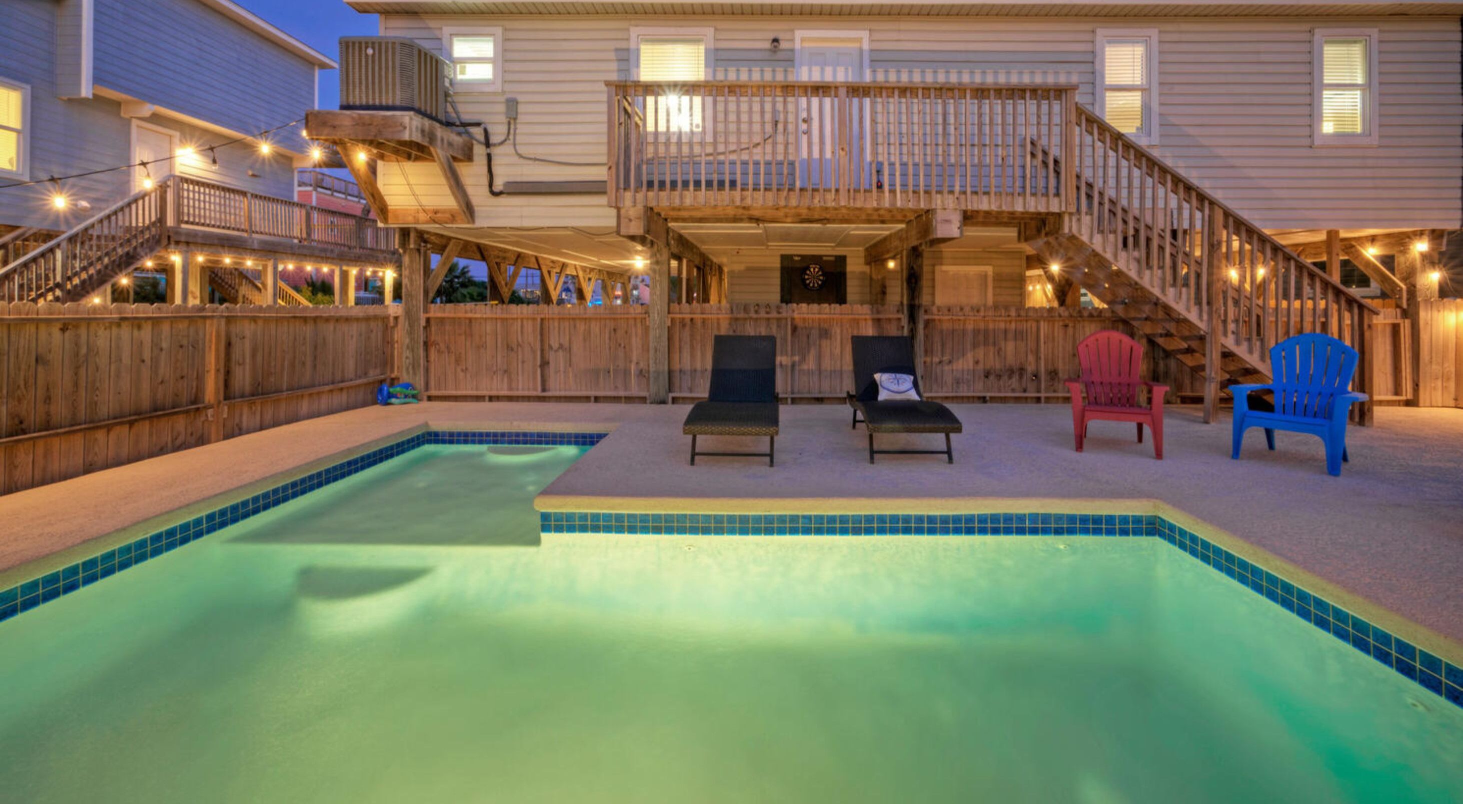 A swimming pool with lounge chairs and a deck area, surrounded by wooden fencing and a house with a balcony.