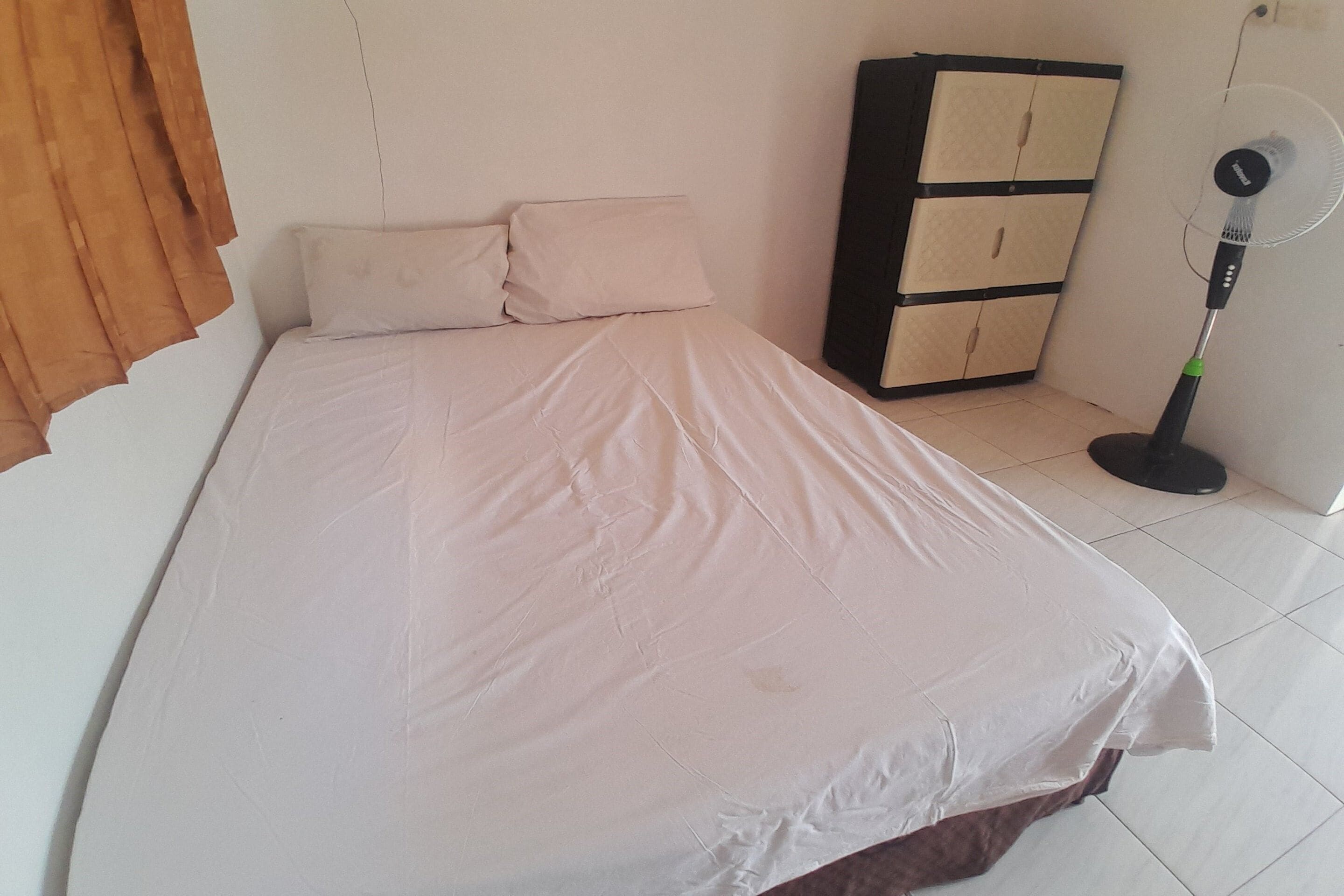 A single bed with a white mattress and two pillows, a standing fan, and a wooden cabinet against a plain wall.