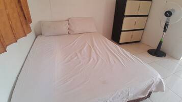 A single bed with a white mattress and two pillows, a standing fan, and a wooden cabinet against a plain wall.