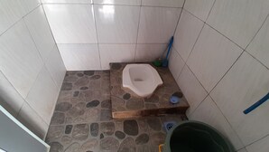 Une salle de bain avec un sol en pierre, des toilettes, un seau vert et un balai.