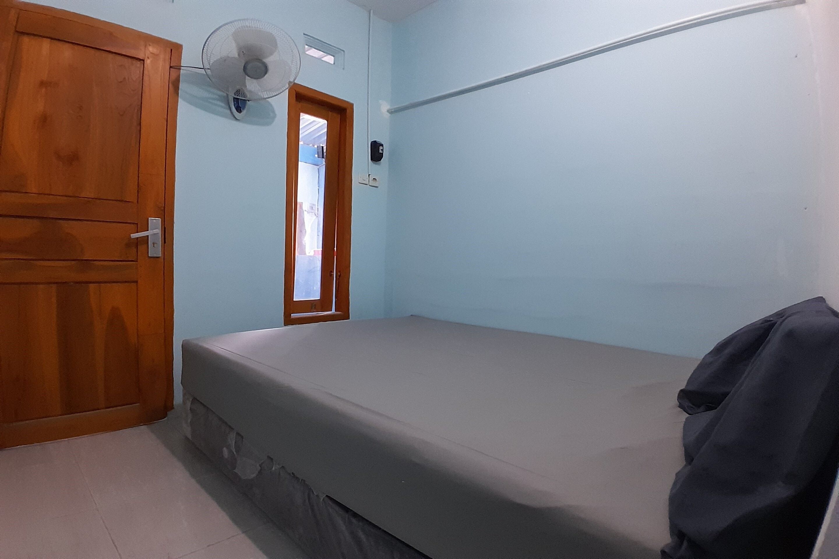A small, single-bed room with a wooden wardrobe, a ceiling fan, and a window with a view of the outside.