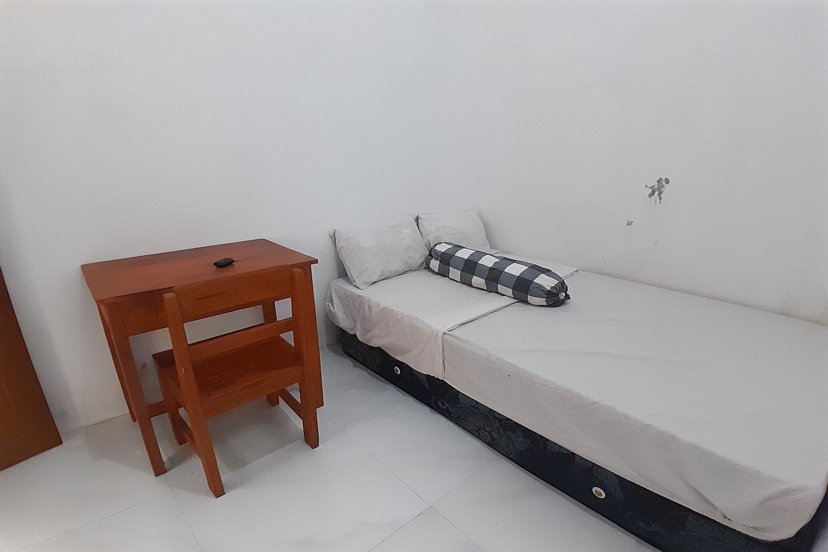Una habitación pequeña con una mesa de madera, una cama con almohada de cuadros y una pared blanca lisa.