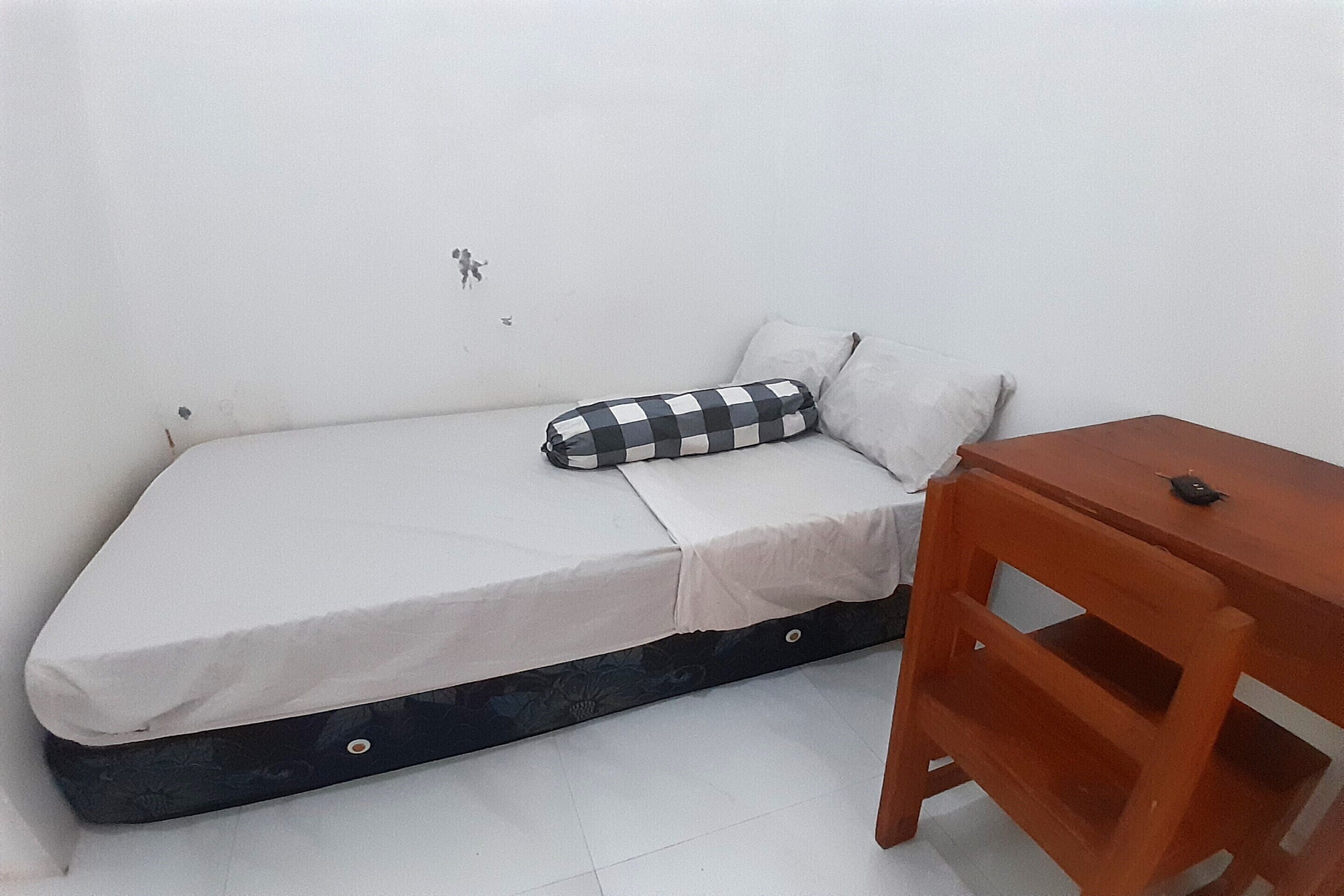 Een kleine eenpersoonskamer met een matras, kussens en een houten nachtkastje.