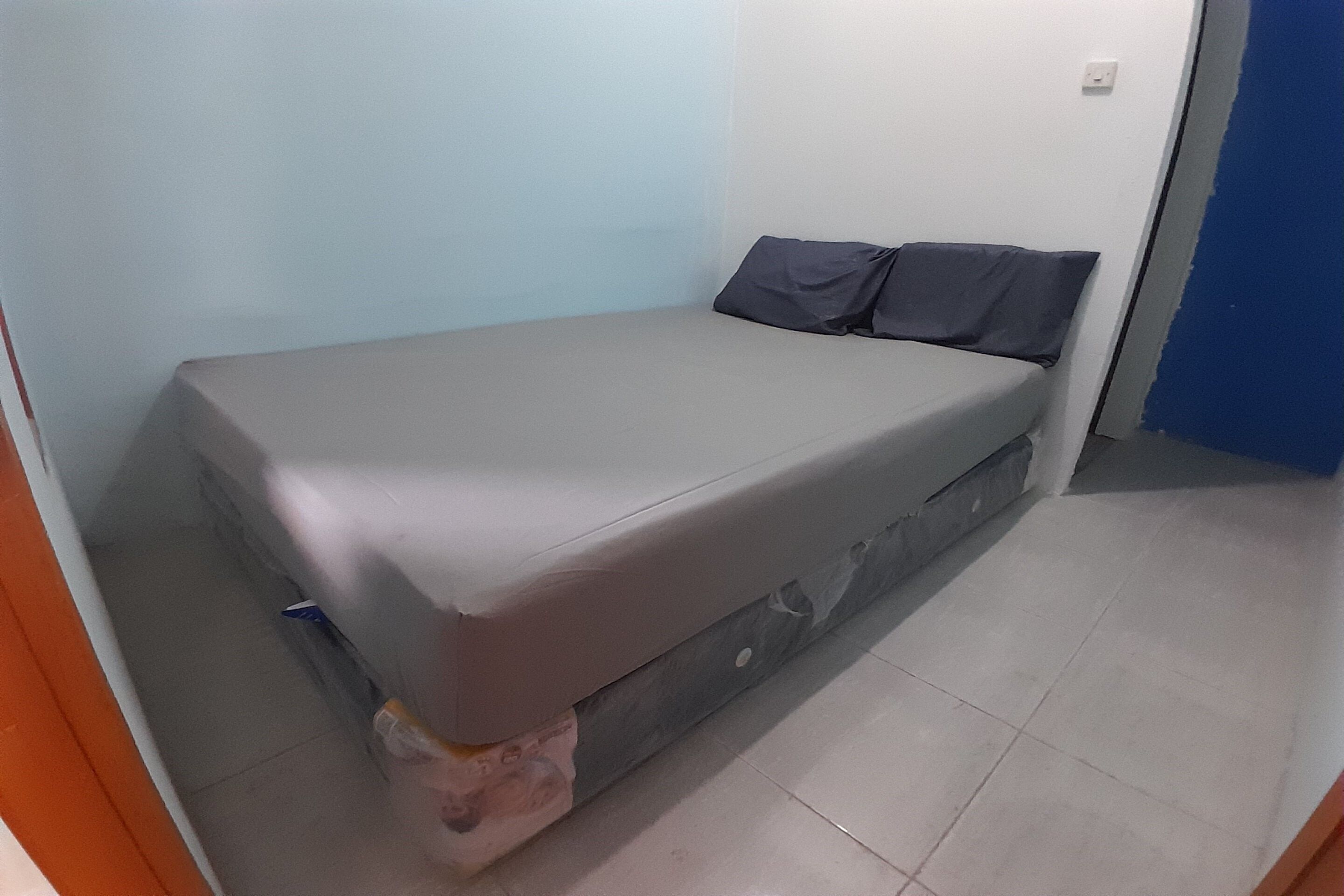 Une petite chambre avec un lit simple, un matelas gris, un oreiller et une couverture.