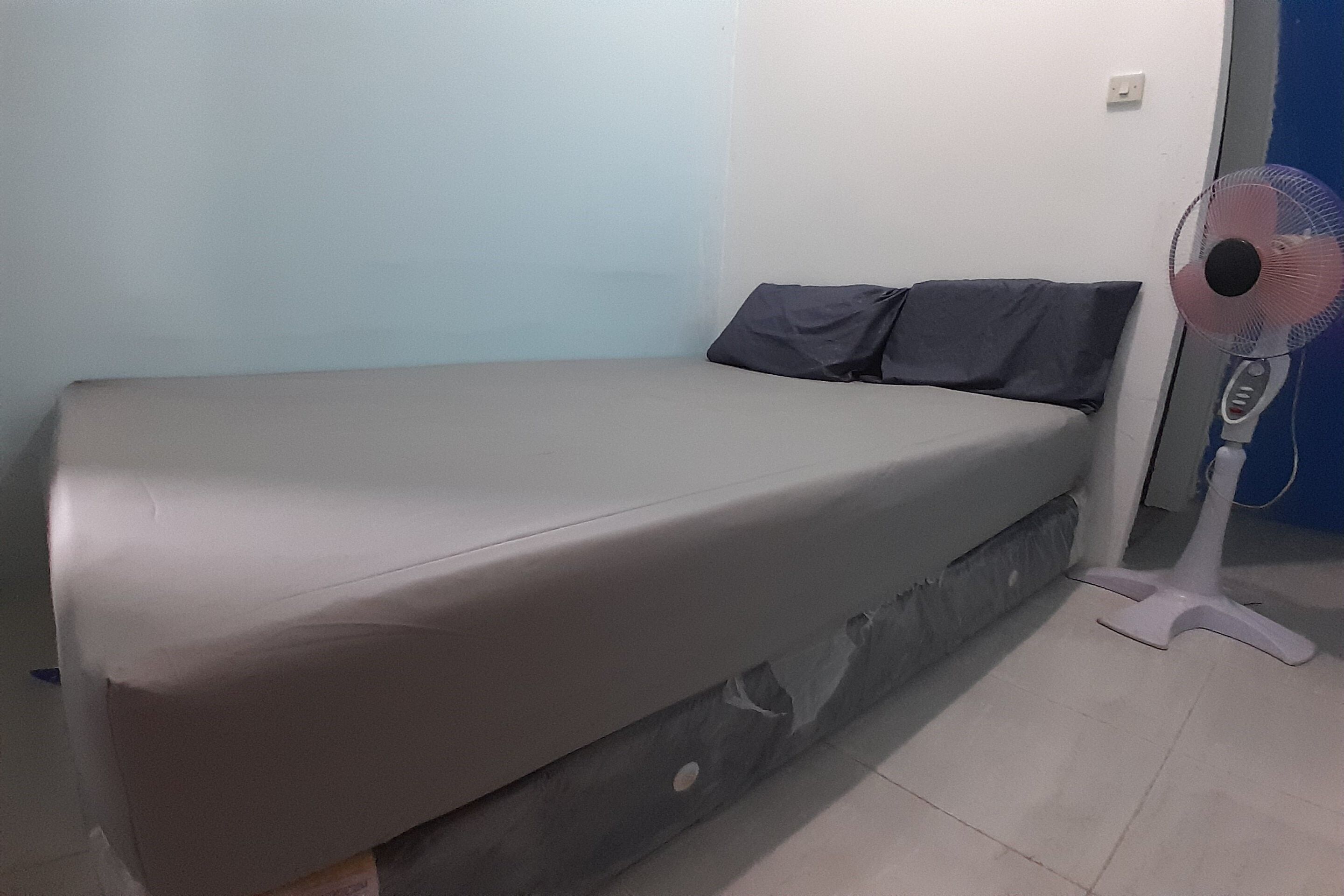 A single bed with a grey mattress and dark pillows, a standing fan, and a tiled floor.