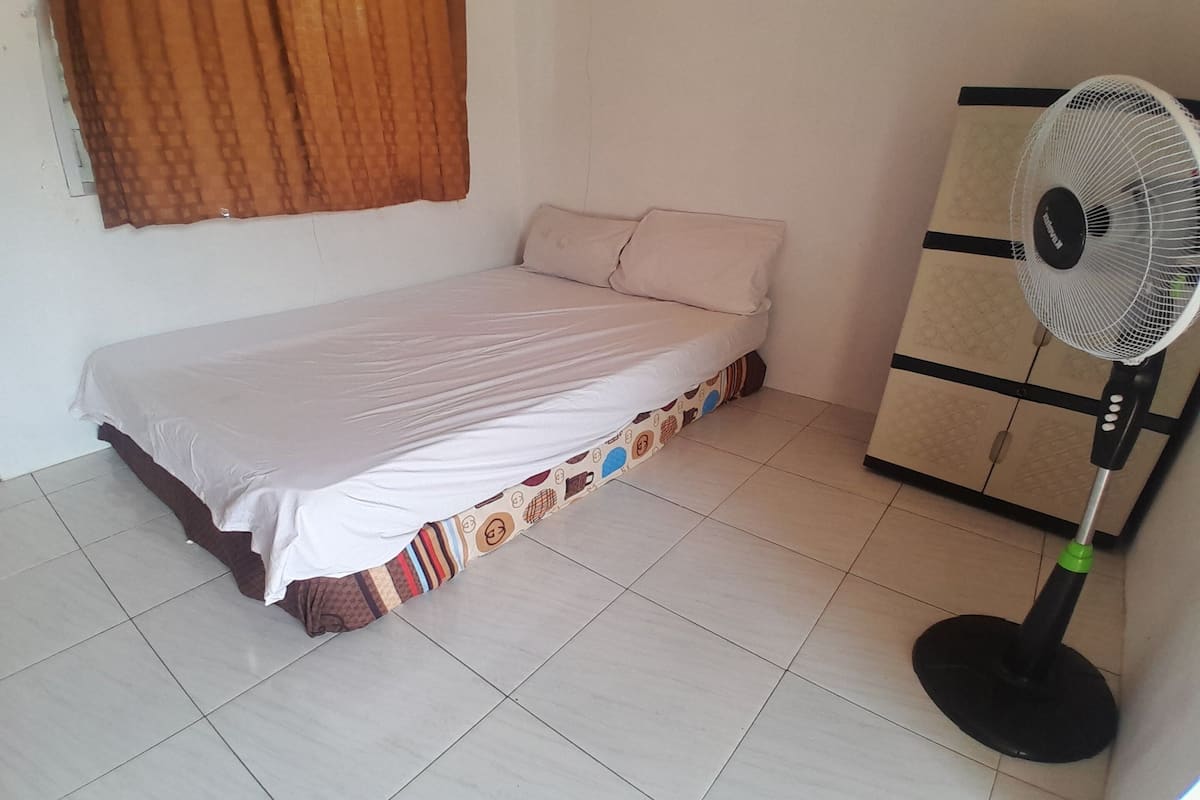 A single bed with a patterned bed skirt, a standing fan, and a cabinet against the wall.