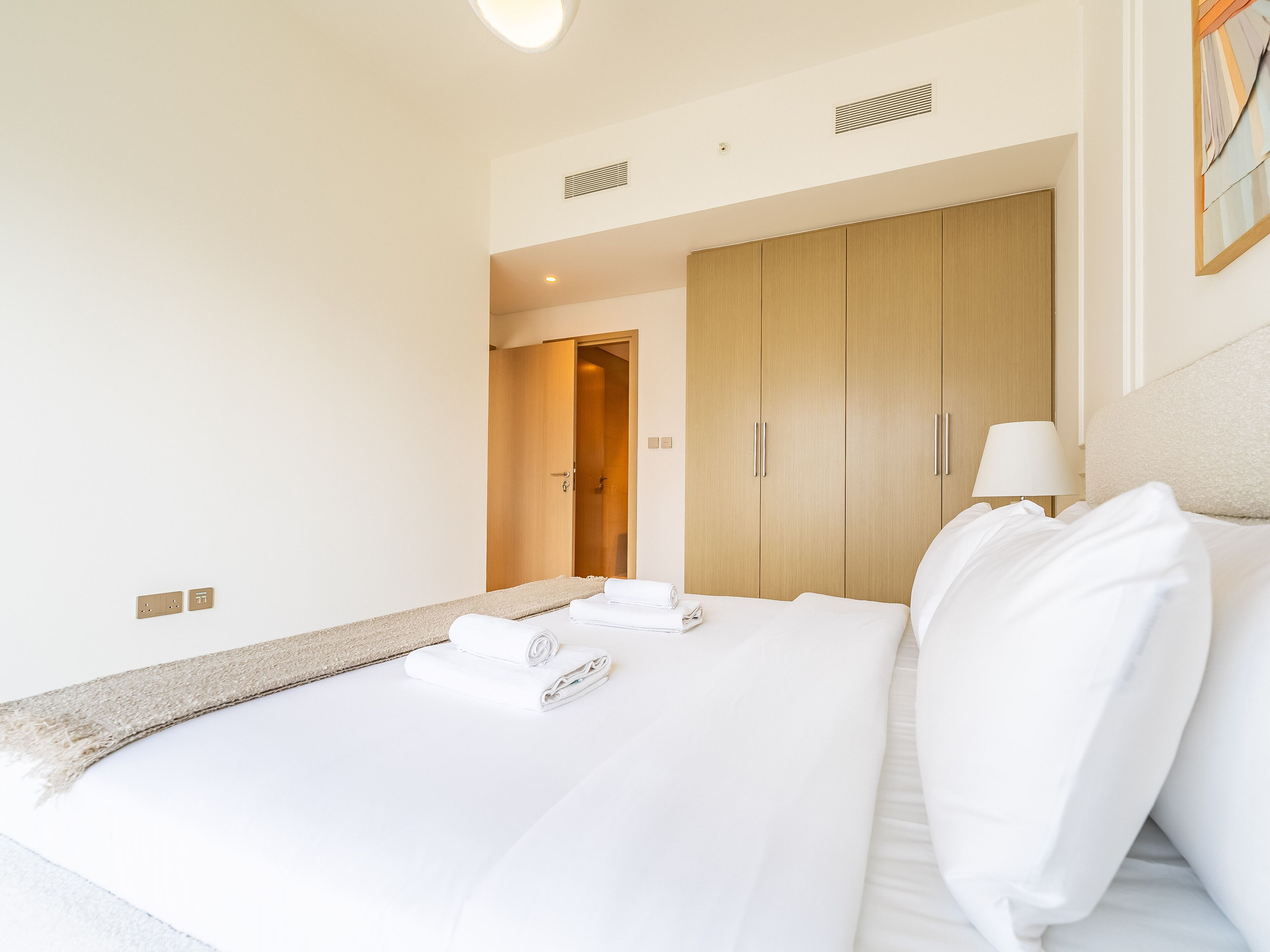 A hotel room with a large bed, a wooden wardrobe, a bedside lamp, and a view of the hallway.