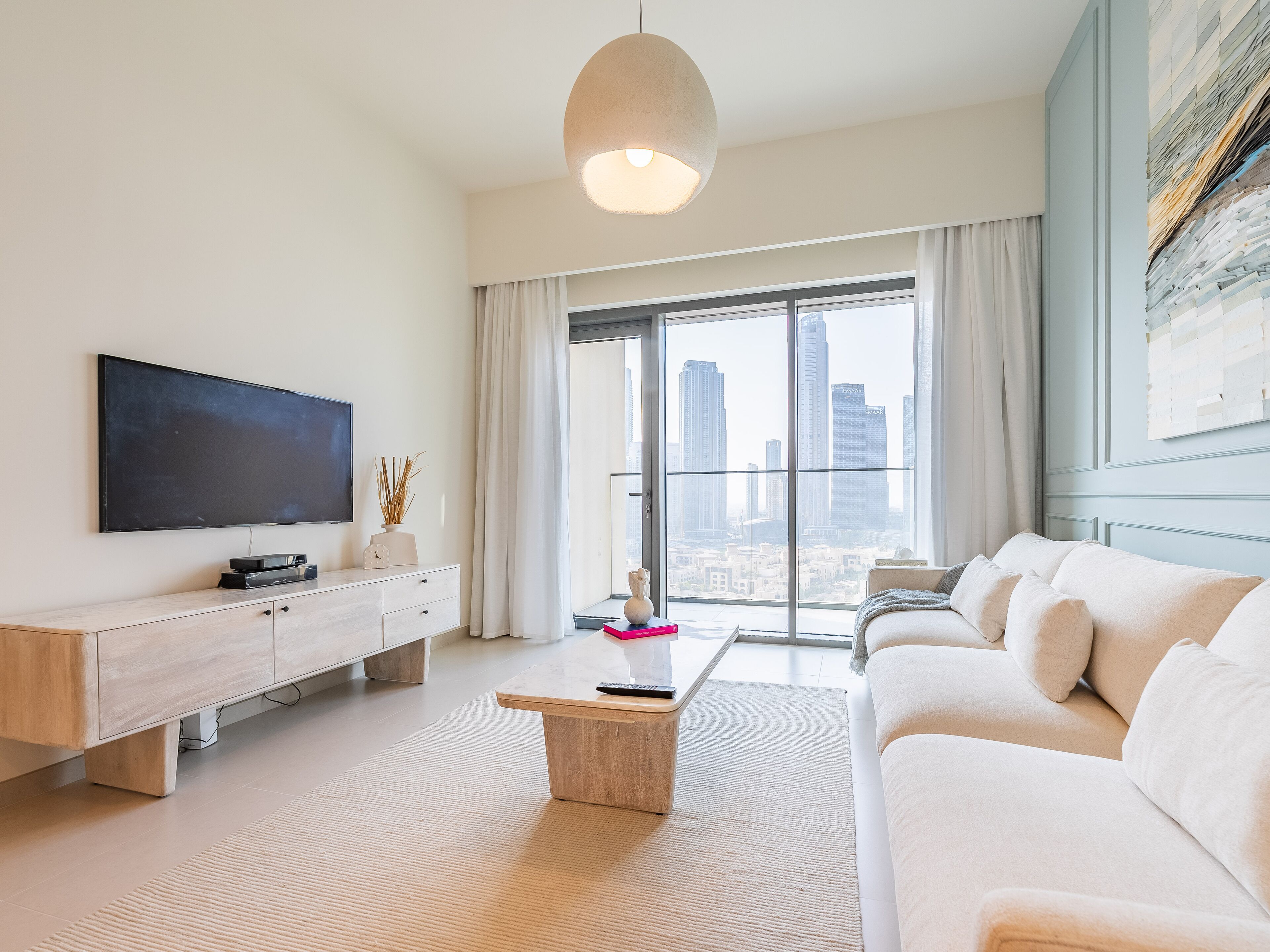 Een moderne woonkamer met een groot flatscreen-televisie, een houten salontafel, een beige bank en een uitzicht op een stadsgezicht door middel van kamerhoge ramen.