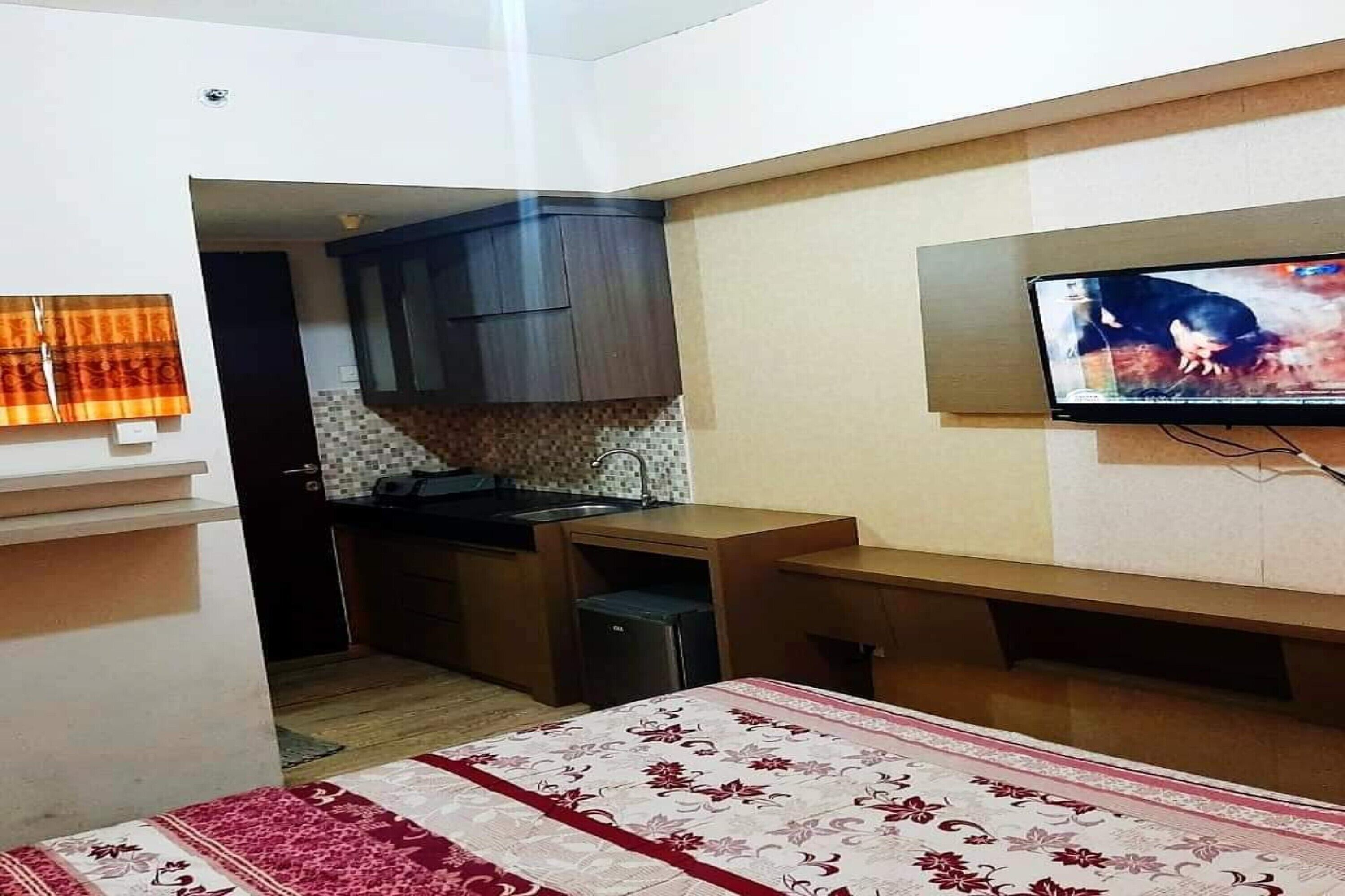 A compact room with a bed, a TV mounted on the wall, a small kitchenette, and a bathroom visible through an open door.