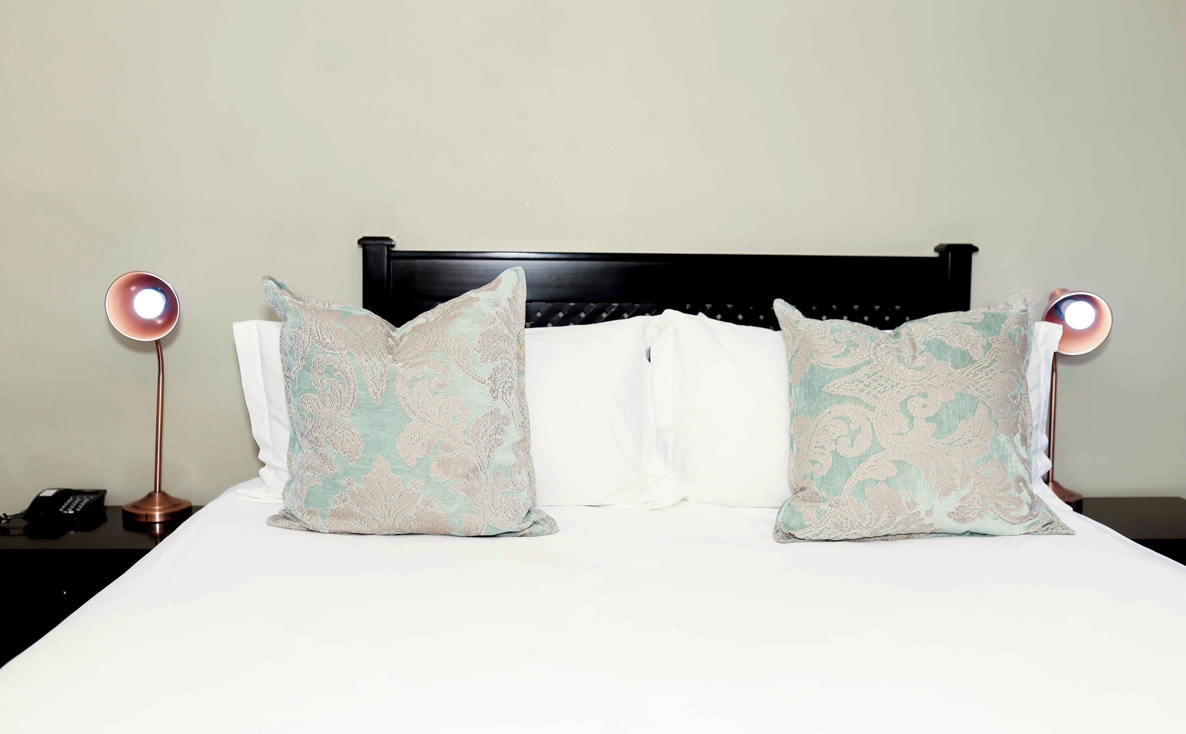 Una cama con dos almohadones decorativos, una lámpara de noche y un teléfono sobre la mesita de noche.