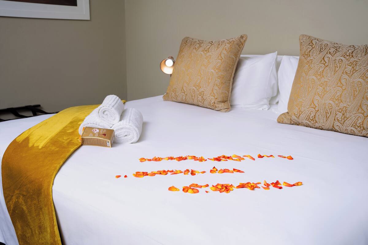 A neatly made bed with a rolled white towel, a box of toiletries, and scattered rose petals.