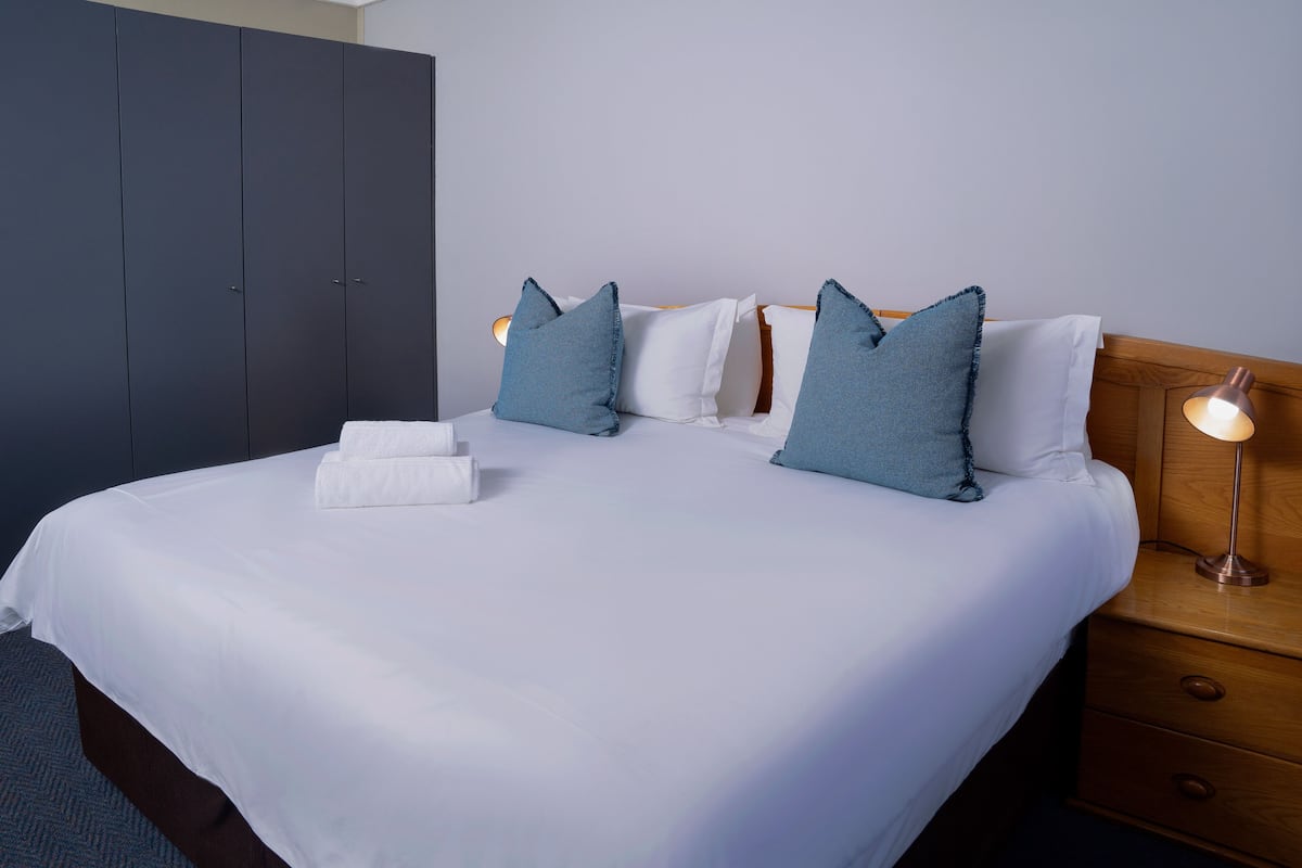 A neatly made bed with white linens and blue pillows, a wooden nightstand with a lamp, and a dark wardrobe.