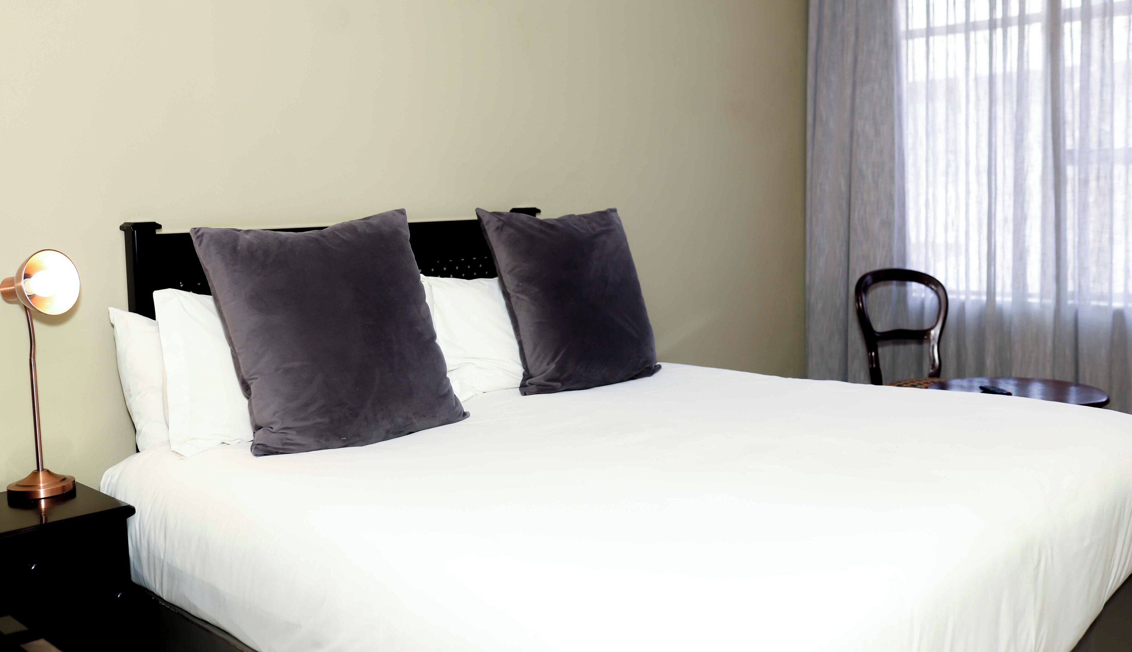 Habitación de hotel con una cama, dos almohadas, una mesita de noche con lámpara y una silla.