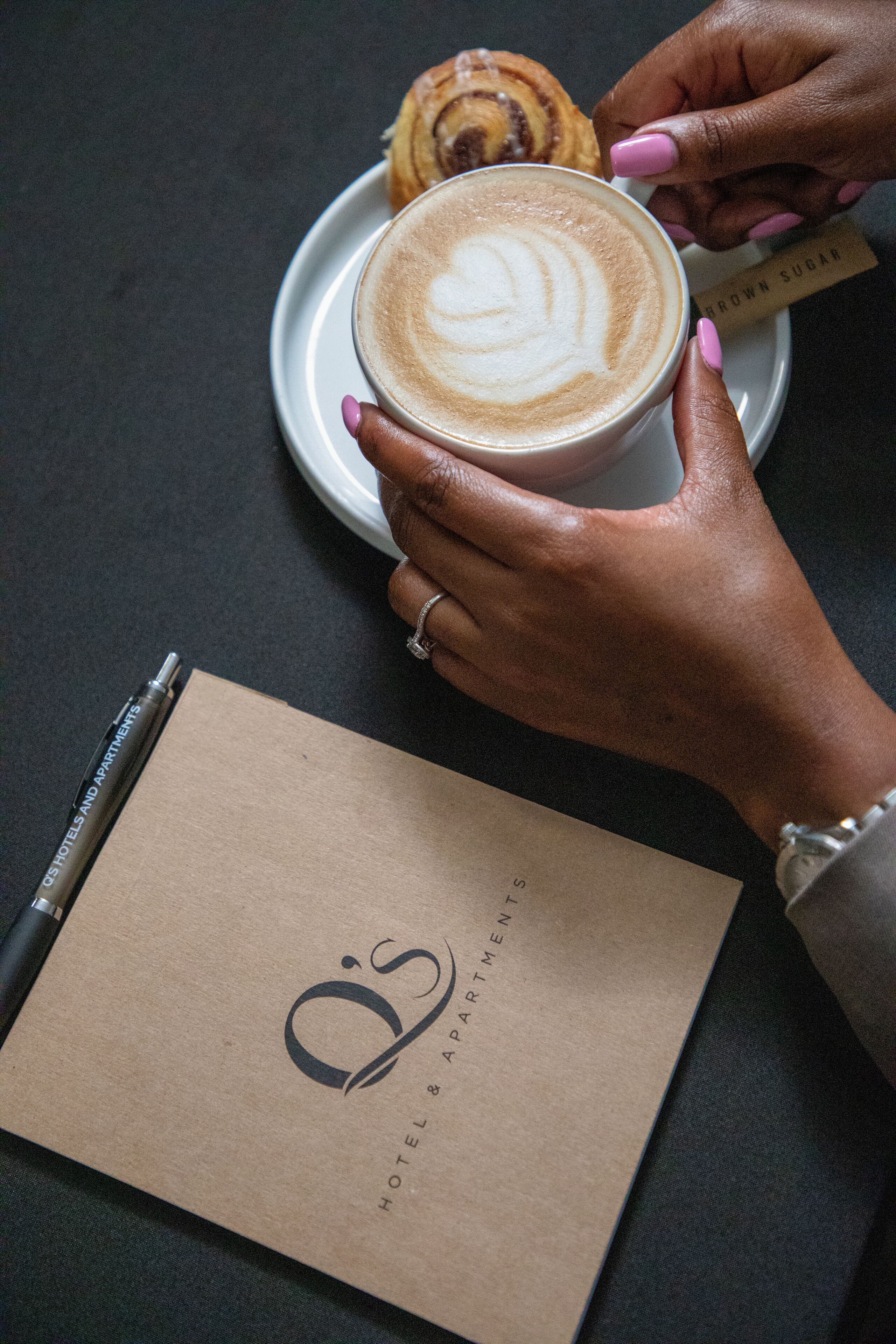 Une main tenant une tasse de café surmontée d’un motif en forme de cœur réalisé avec de la mousse de lait, une pâtisserie sur une assiette et une carte de visite portant l’inscription « O’S HOTEL & APARTMENTS » sur une table.