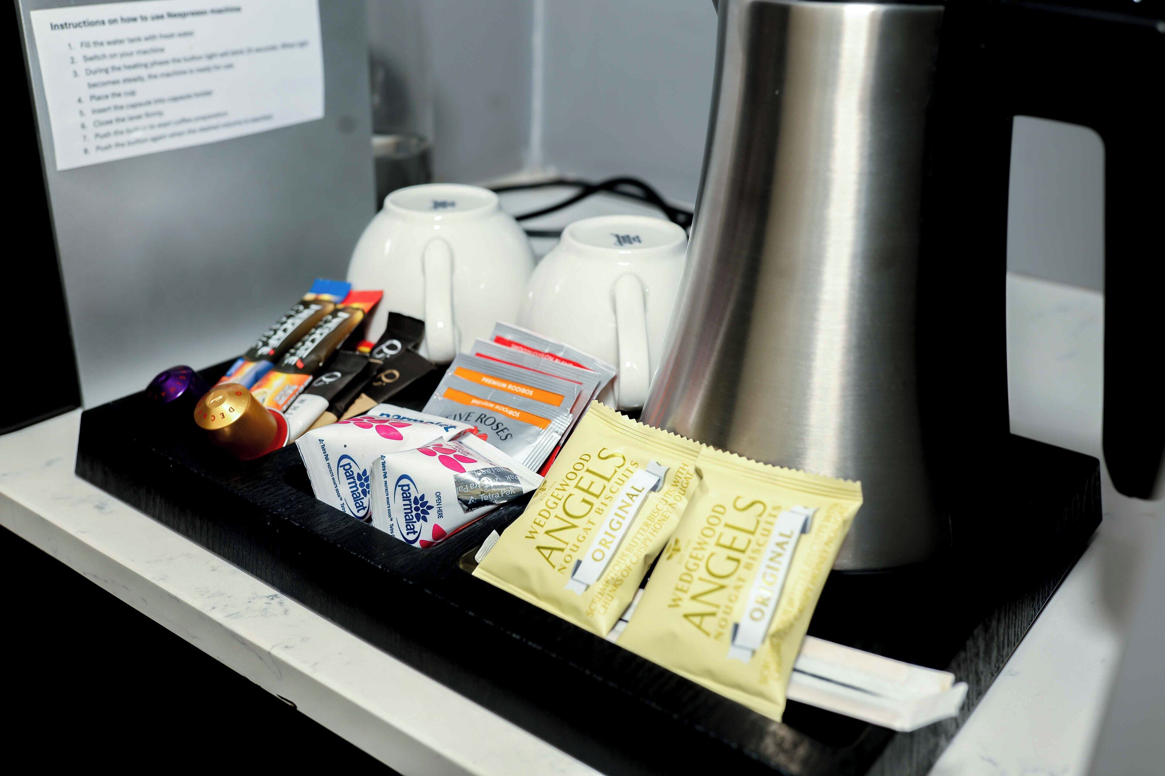 Una bandeja con diversos objetos, como un control remoto, paquetes de aperitivos y una cafetera.