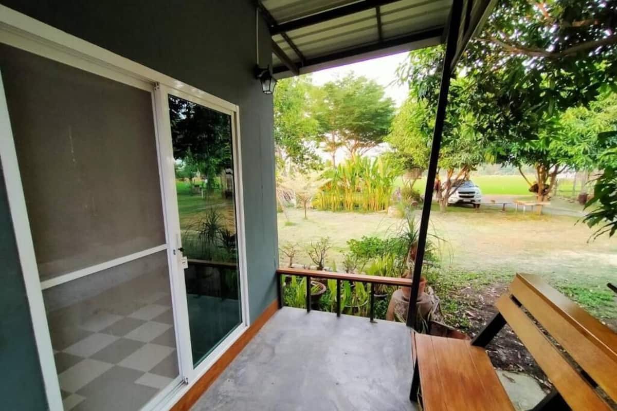 Ein überdachter Balkon mit Betonboden, einer hölzernen Bank und Blick auf einen Garten mit verschiedenen Pflanzen und einem parkenden Auto.