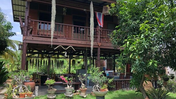 A traditional wooden building with a balcony, surrounded by lush greenery and potted plants. - Metha Country View Homestay Singburi (Khai Bang Rachan)