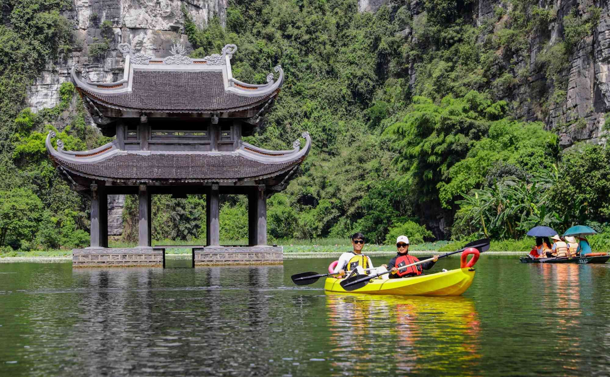 A traditional pavilion with a tiled roof and ornate eaves, situated by a calm lake with kayakers.