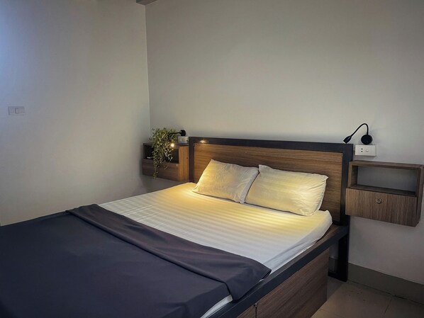 A neatly made bed with a wooden headboard, a bedside table with a lamp, and a wall-mounted power outlet. - Hanoi EcoStay 2 hostel (Hanoi)