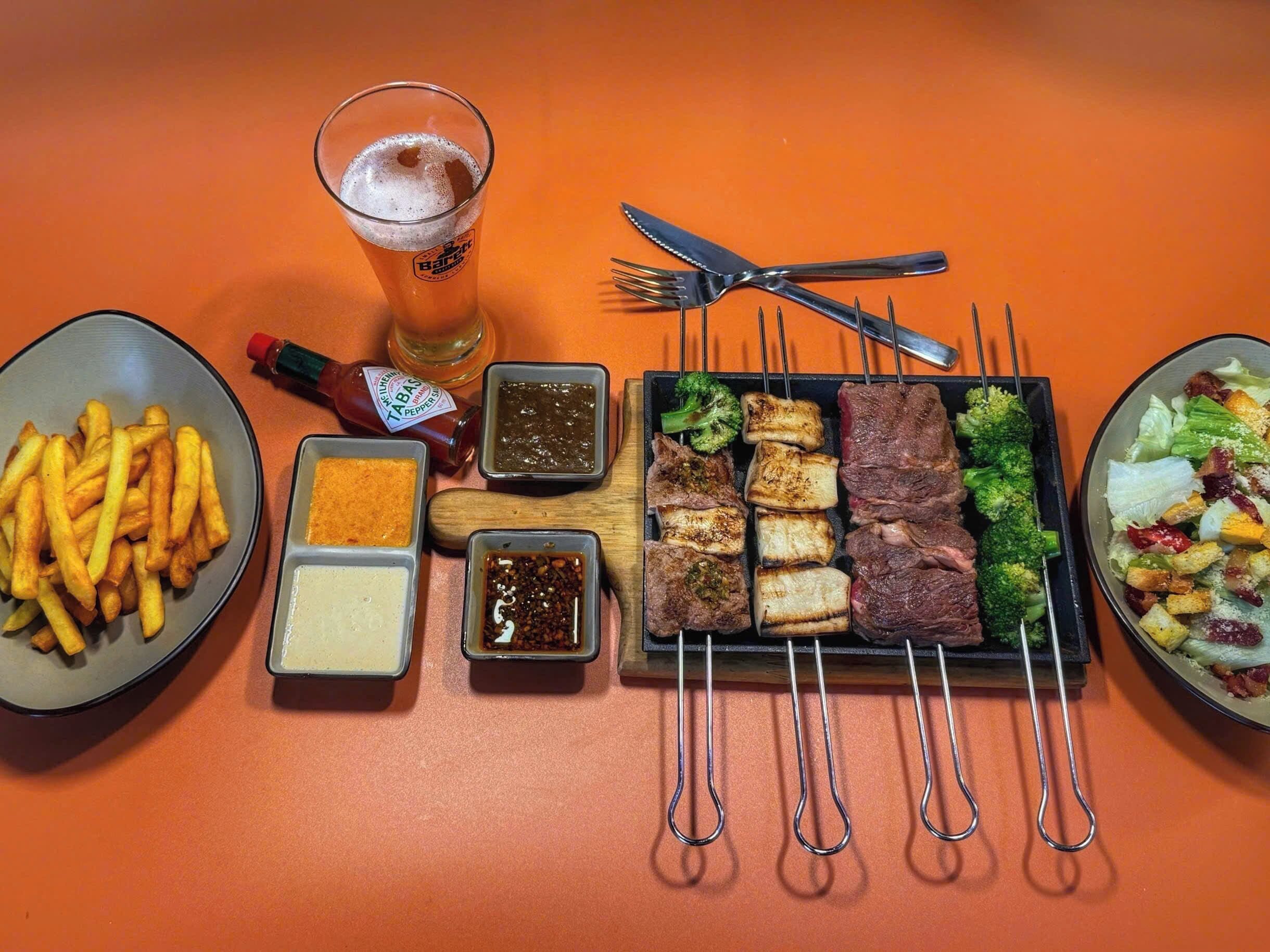A meal with various dishes and condiments, including fries, grilled meats, and vegetables, on an orange table.