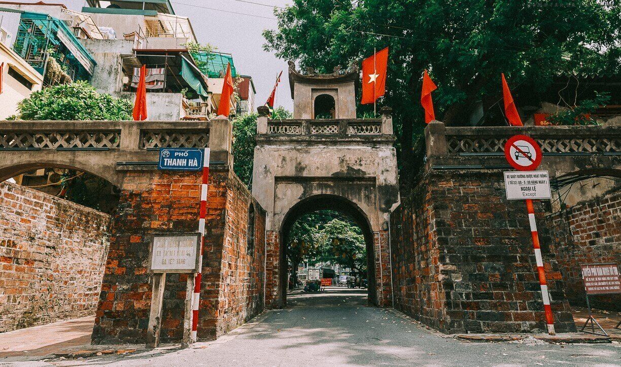 A historic stone archway with flags, a sign indicating a no-entry for vehicles, and a street leading through it.