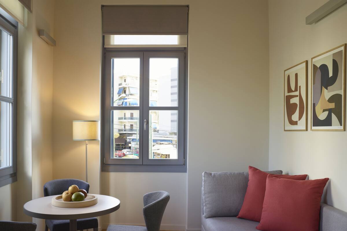 A living room with a gray sofa, a small round table with fruit, a chair, a window with a view of buildings, and abstract art on the wall.