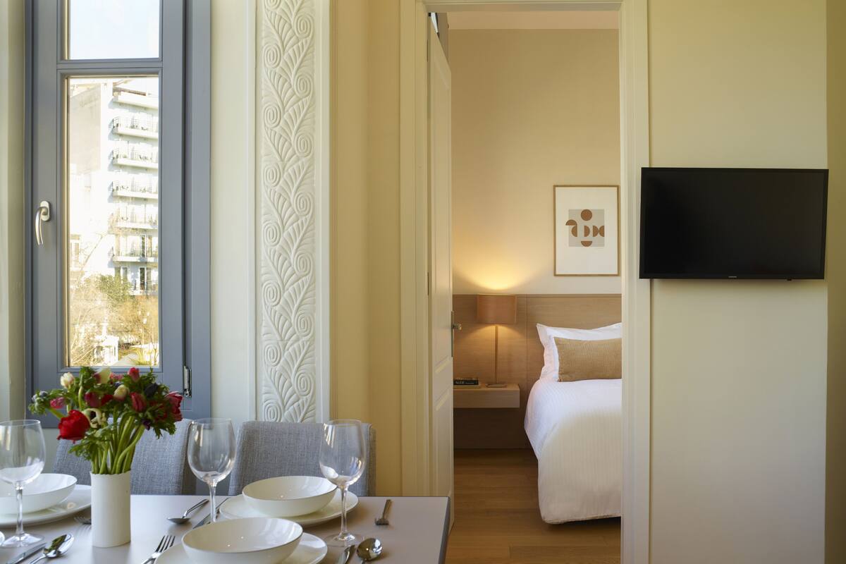 A hotel room with a dining area, a bed, a TV, and a view of buildings outside.