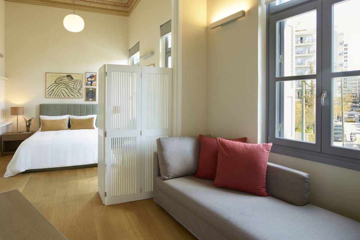 A hotel room with a bed, a sofa, a wardrobe, and a window with a city view.