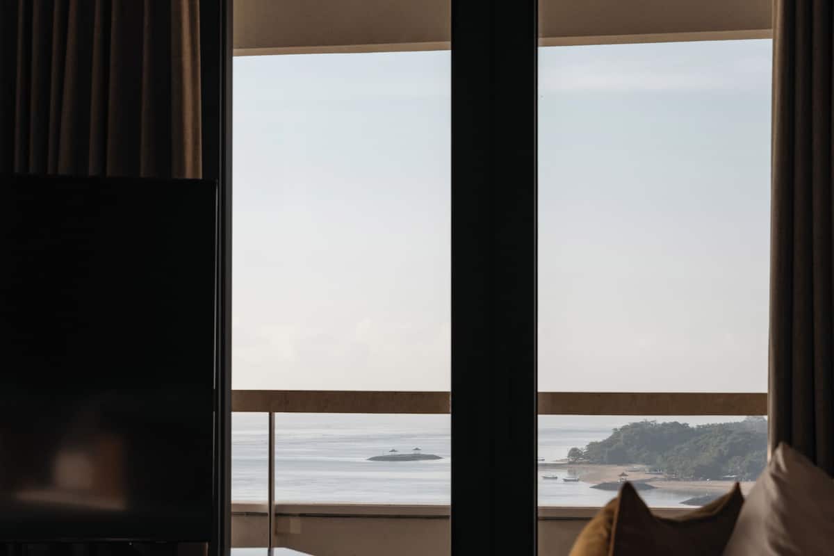 A hotel room with a bed, a TV, and a large window offering a view of the sea and a distant island.