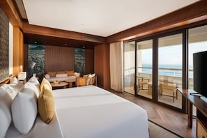 Una habitación de hotel con una cama grande, un sofá, una mesa de comedor y un televisor. La habitación tiene un ventanal con vista al océano.