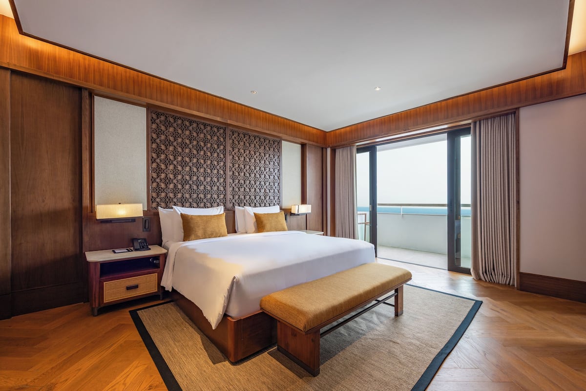 A spacious hotel room with a large bed, a bench, a nightstand, and a view of the sea through a large window.