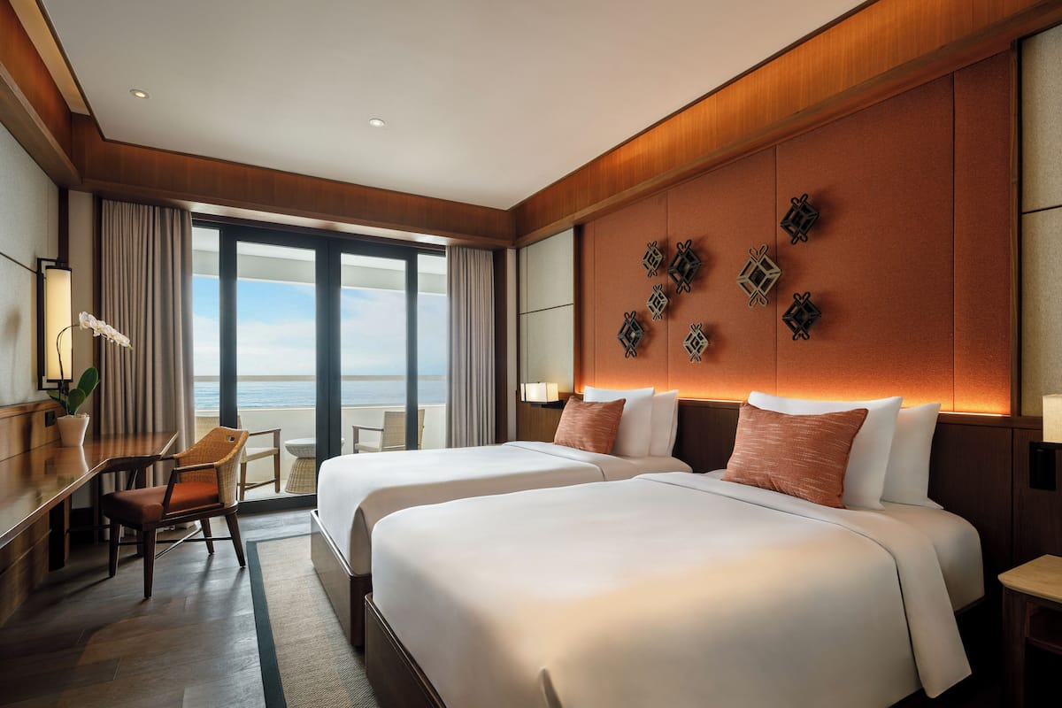 A hotel room with two beds, a desk, a chair, and a view of the ocean.